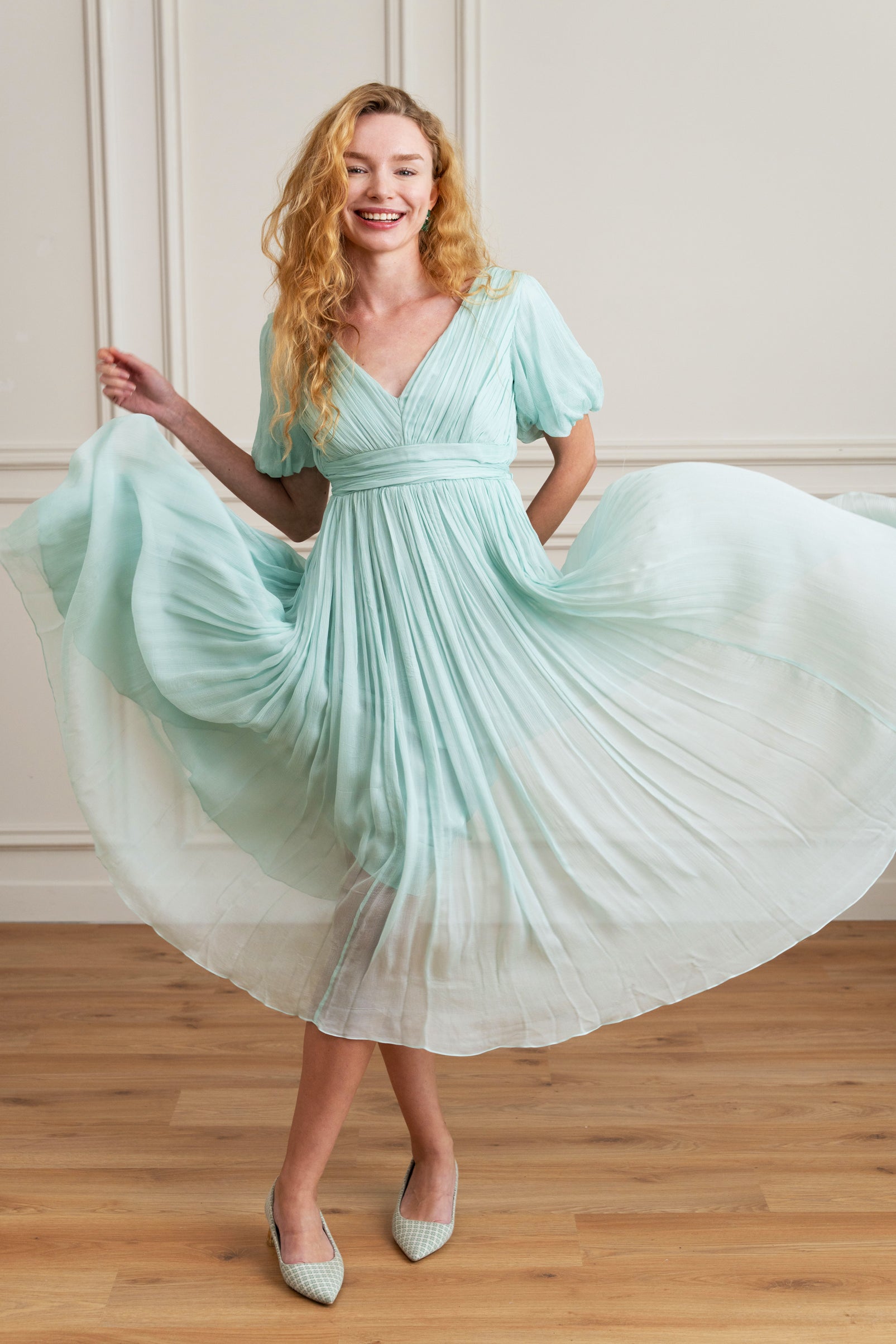 Woman in a light BLUE dress twirling in a room with wooden flooring and beige walls.