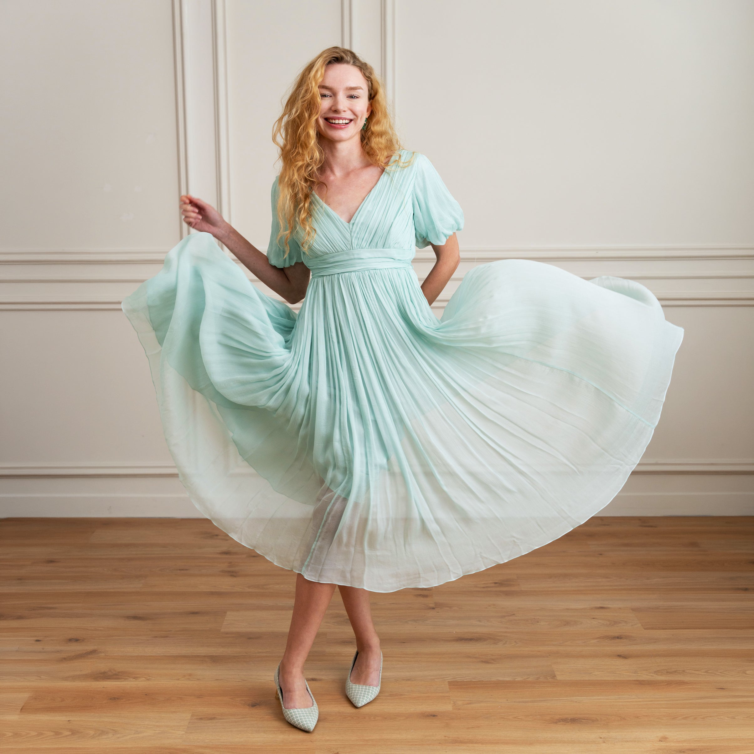 Woman in a light BLUE dress twirling in a room with wooden flooring and beige walls.