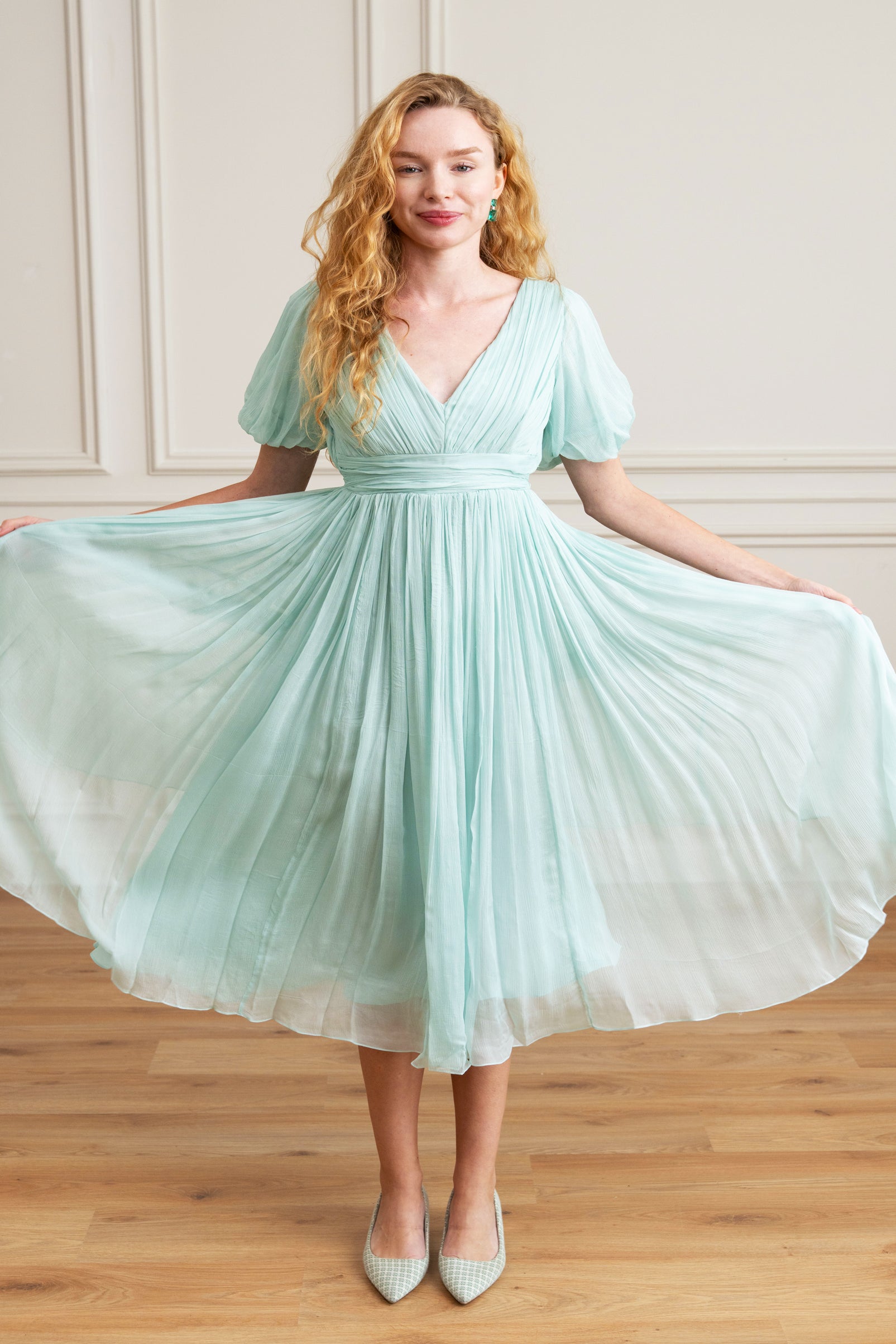 Woman wearing a light blue dress in a room with wooden flooring and white paneled walls.