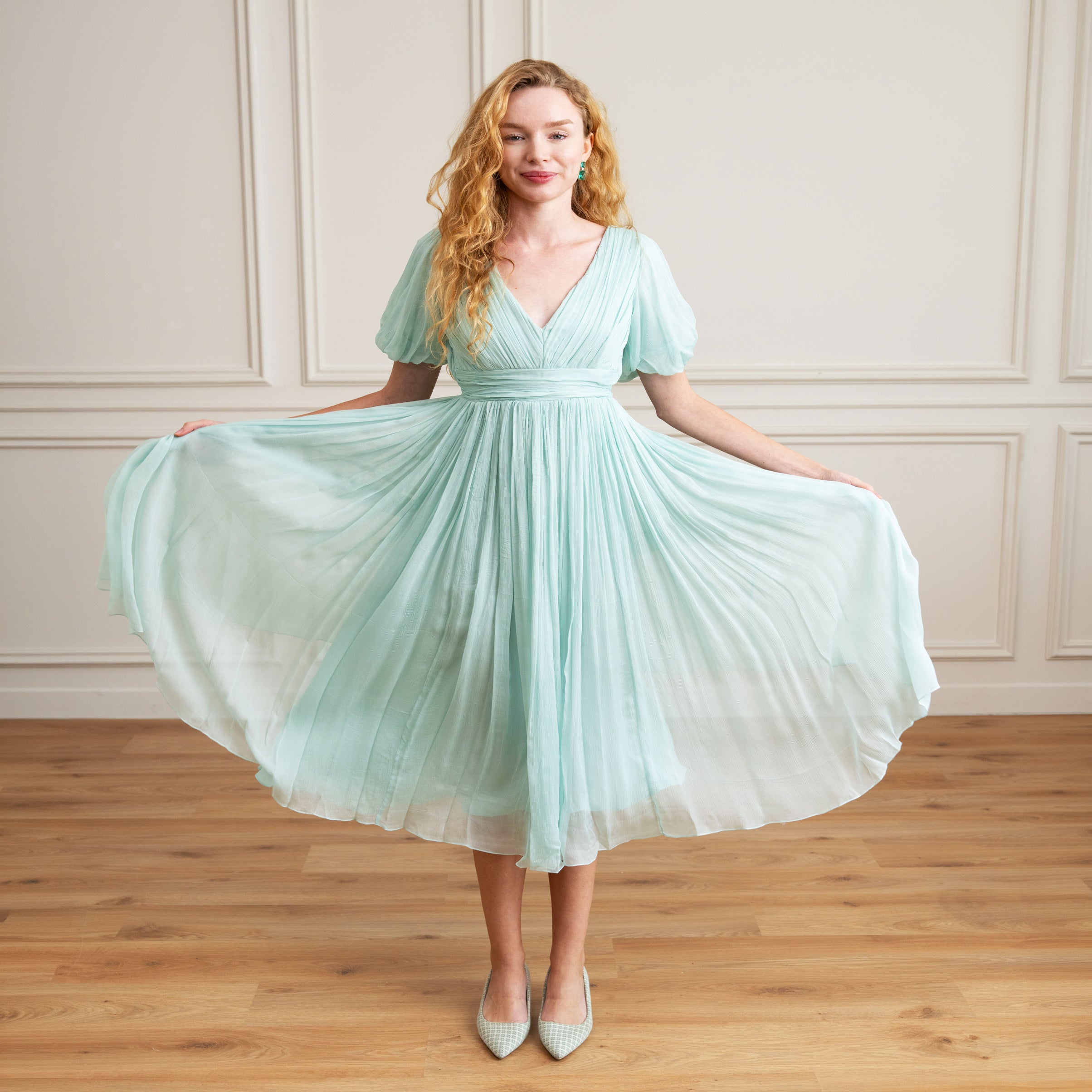 Woman wearing a light blue dress in a room with wooden flooring and white paneled walls.