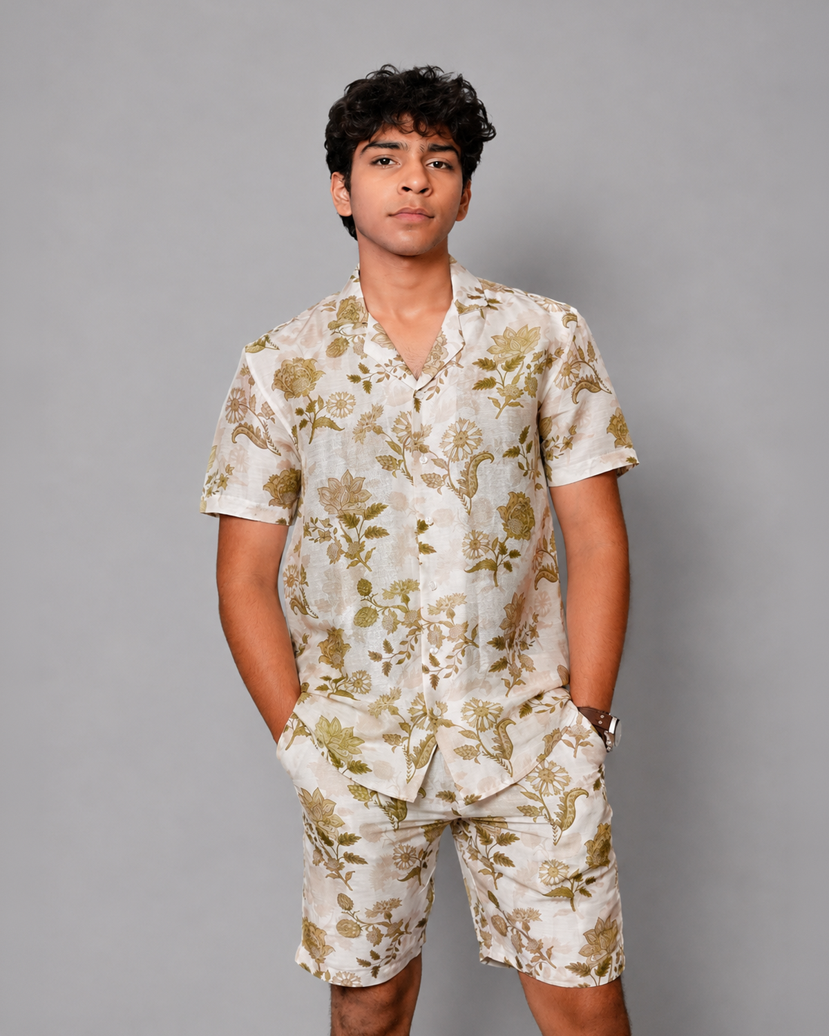 A young man modeling a matching short-sleeve button-up shirt and shorts set in a light cream color with a muted olive-green floral pattern, set against a solid gray studio background.