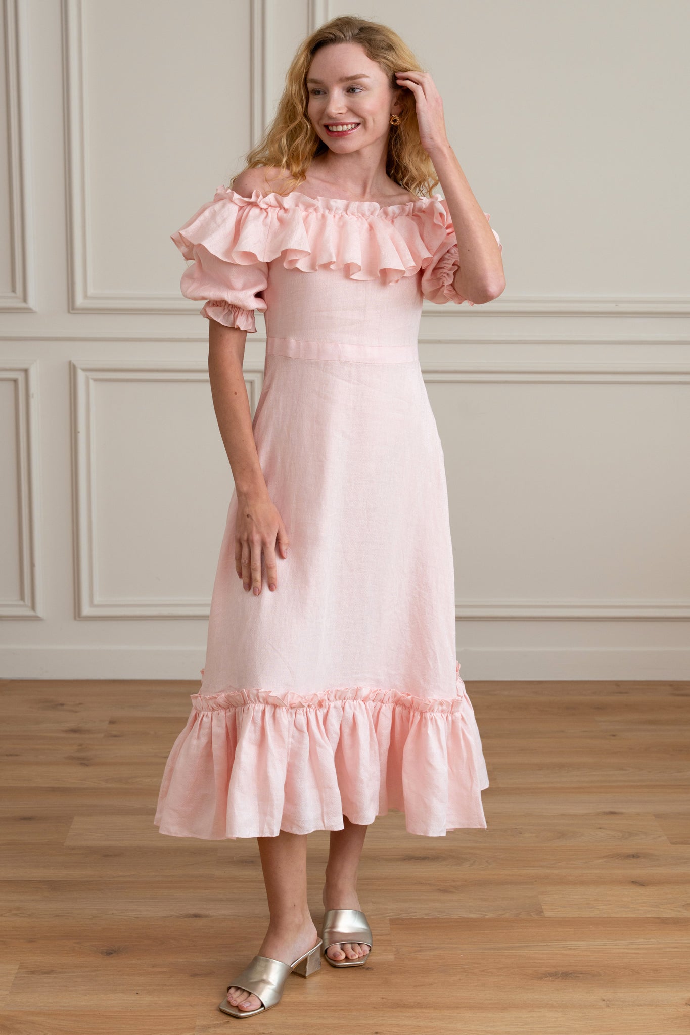 Woman wearing a pink off-shoulder dress with ruffled details in a room with wooden flooring and paneled walls.