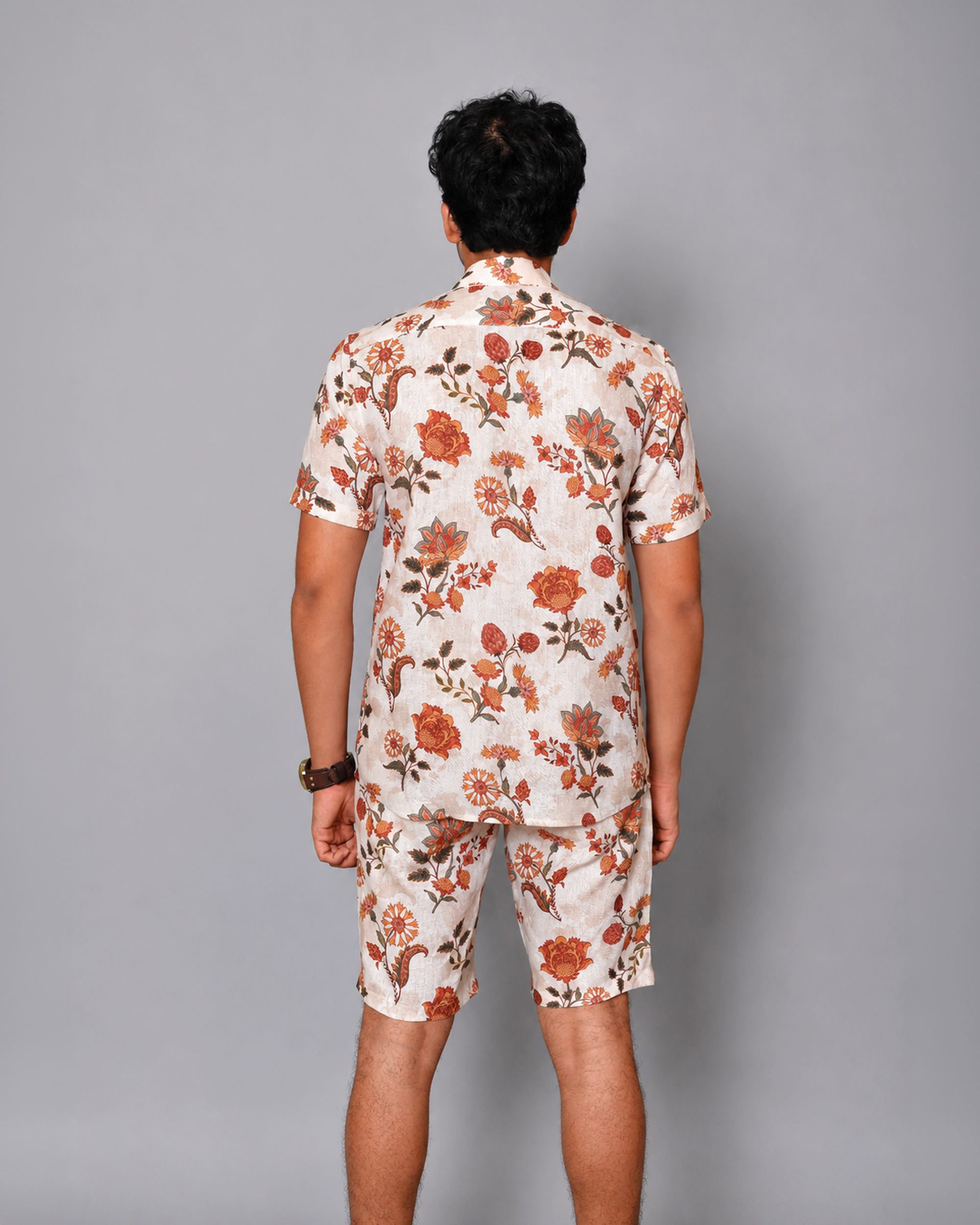 A man viewed from behind wearing a matching short-sleeve button-up shirt and shorts set in a cream base fabric with a rust-orange and brown floral print, against a solid gray studio background.