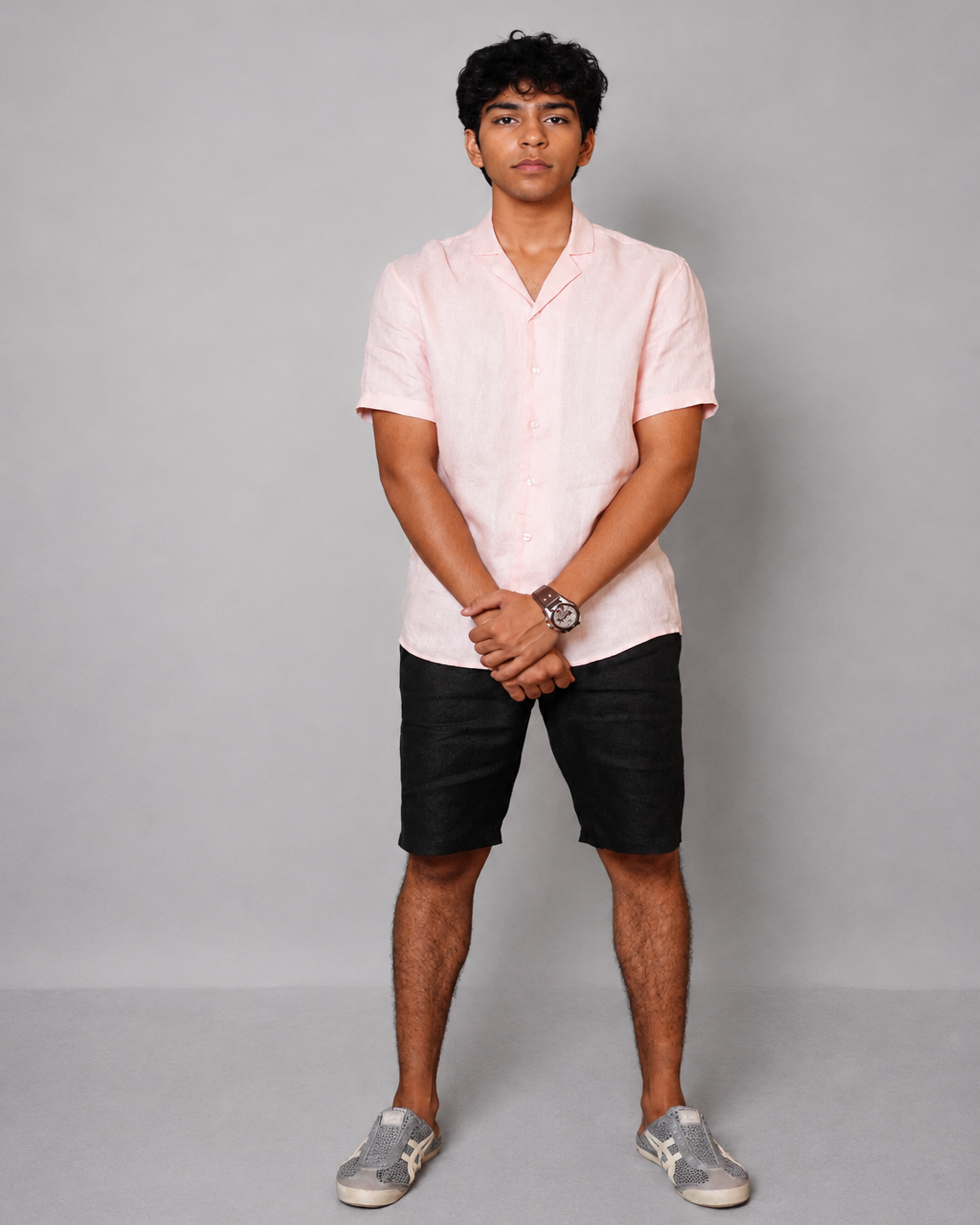 A man modeling a light pink short-sleeve button-up shirt, black shorts, and gray slip-on sneakers against a solid gray studio background.