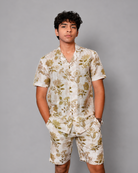 A young man modeling a matching short-sleeve button-up shirt and shorts set in a light cream color with a muted olive-green floral pattern, set against a solid gray studio background.