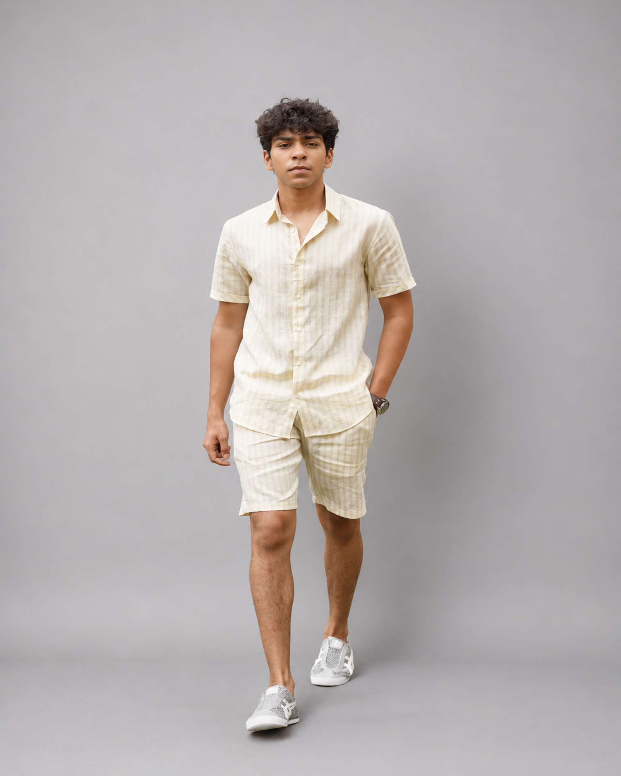 A man modeling a matching short-sleeve button-up shirt and shorts set in a light cream or off-white fabric with a subtle vertical stripe pattern, paired with white sneakers, against a solid gray studio background.