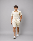 A man modeling a matching short-sleeve button-up shirt and shorts set in a light cream or off-white fabric with a subtle vertical stripe pattern, paired with white sneakers, against a solid gray studio background.
