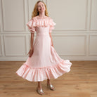 Woman wearing a pink off-shoulder dress with ruffles in a room with wooden floor and white paneled walls.