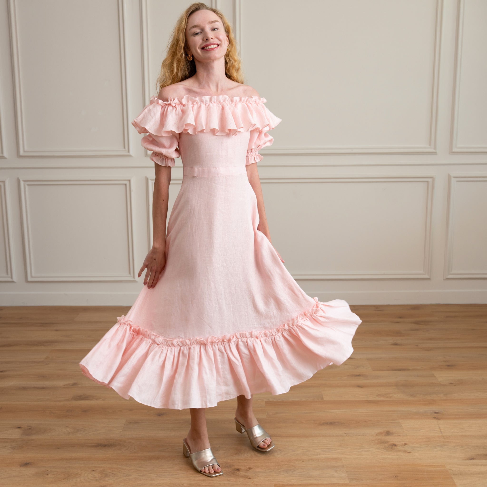 Woman wearing a pink off-shoulder dress with ruffles in a room with wooden floor and white paneled walls.